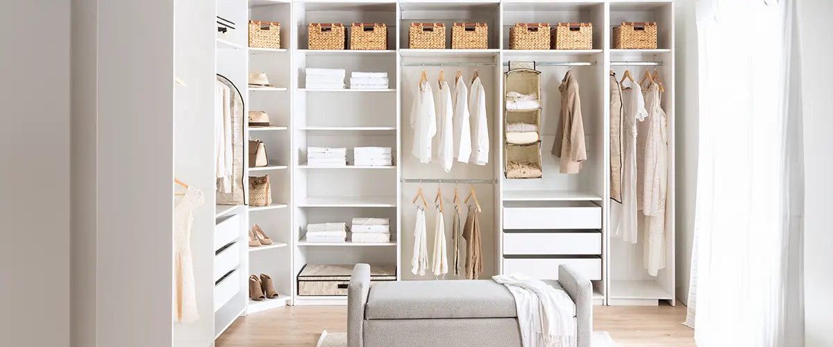 Spacious organized walk-in closet system in Cold Springs, with white shelves, wicker baskets, and neatly arranged clothing, featuring a cozy upholstered bench.