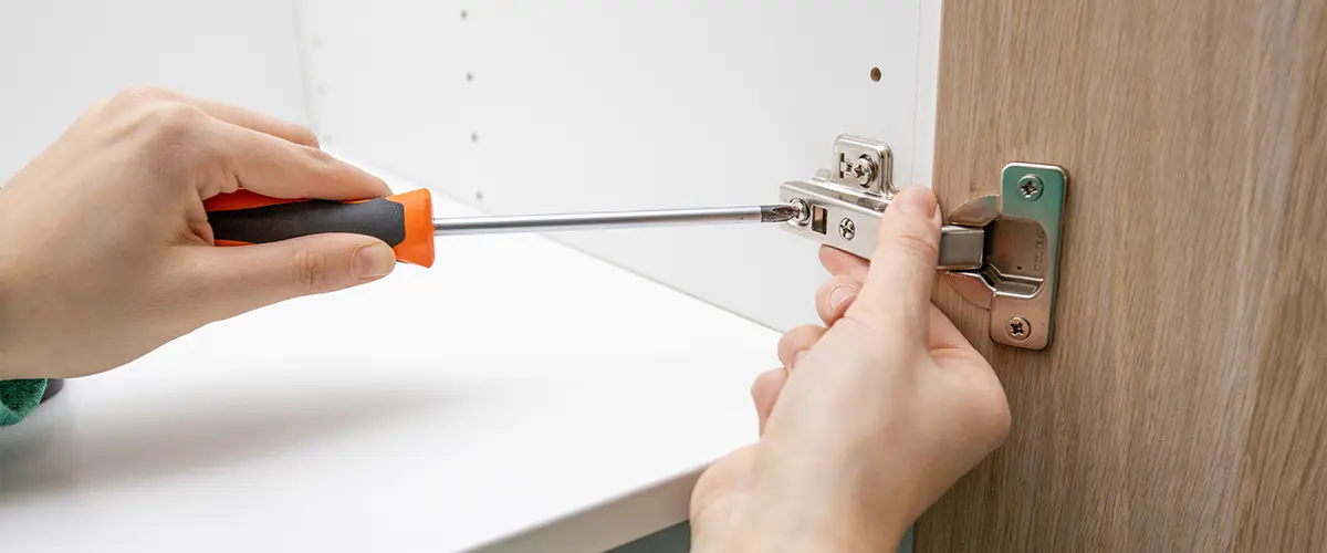 Person using a screwdriver to install a soft-close hinge on a wooden cabinet door in a modern kitchen.